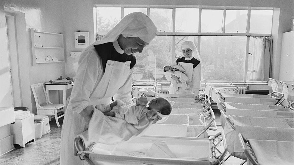 A black and white photo from the 1960s shows wo maternity nurses in full uniform and head-dress comfort babies in a maternity ward, holding them above a row of trolleys