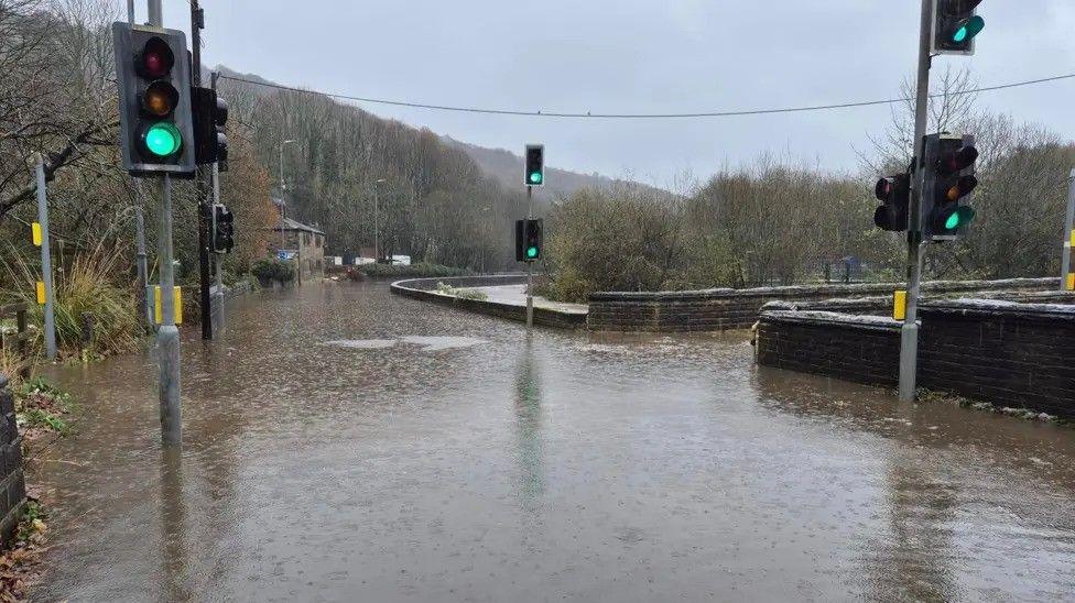 A heavily flooded road with no cars on it. Traffic lights, which are on green, flank the road. The sky is grey and the trees in the background are bare and autumnal.