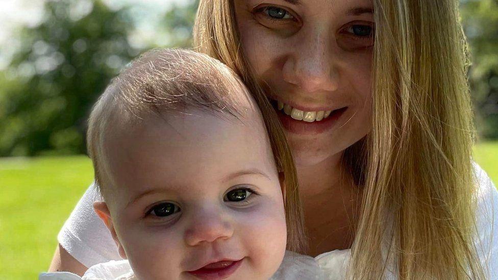 Rebecca Ableman is smiling while holding her daughter, Autumn. They are both outside on the grass during a sunny day and are wearing white. Autumn is smiling and looking directly at the camera. Rebecca is doing the same and has long blonde hair.