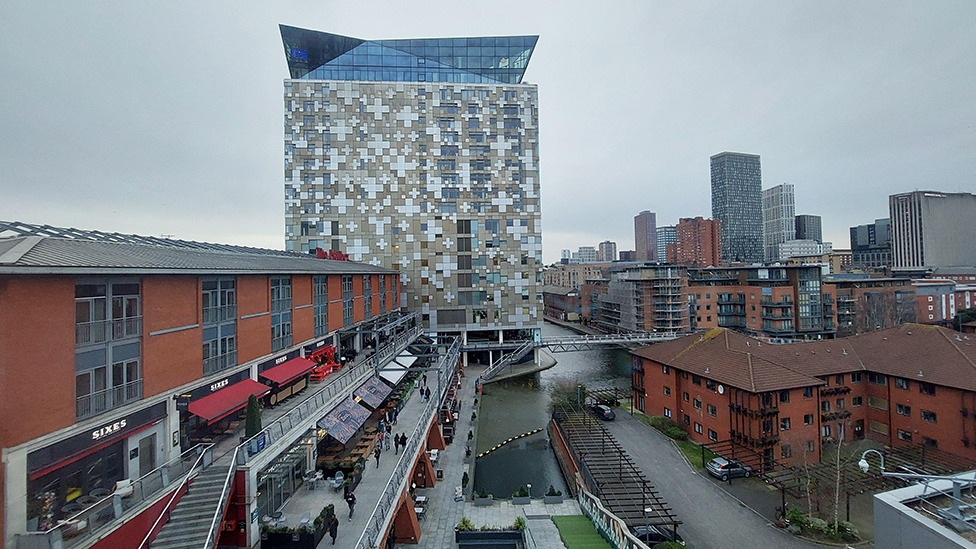 A view of a canal, lined on either side by tall red brick buildings with a number of bars and cafes on the left hand side and a tall metal building in the background