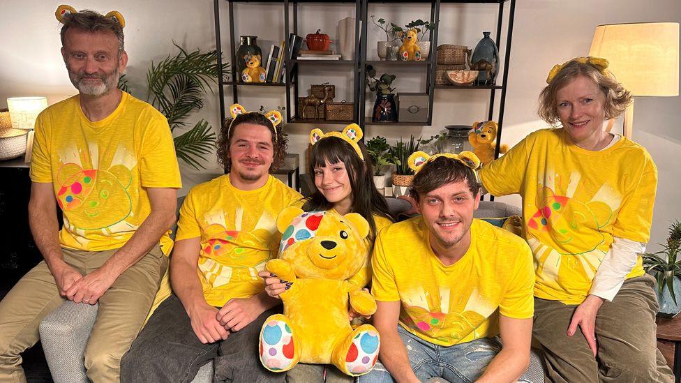 The cast of Outnumbered sitting on a sofa wearing yellow Children In Need T-shirts and Pudsey ears, with a Pudsey cuddly toy