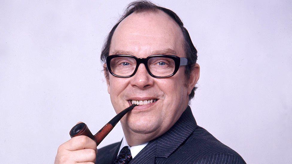 Eric Morecambe holding a pipe between his teeth and smiling. He has thinning dark hair, black-framed glasses and is wearing a navy pinstripe suit and tie.