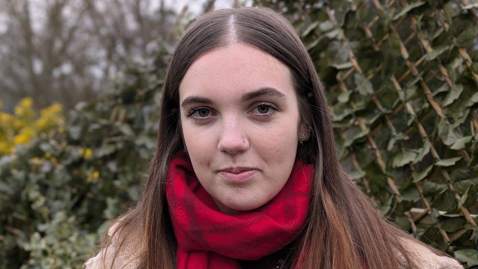 Mollie looks straight ahead, standing in front of a some greenery - she has long brown hair parted in the middle and wears a red scarf