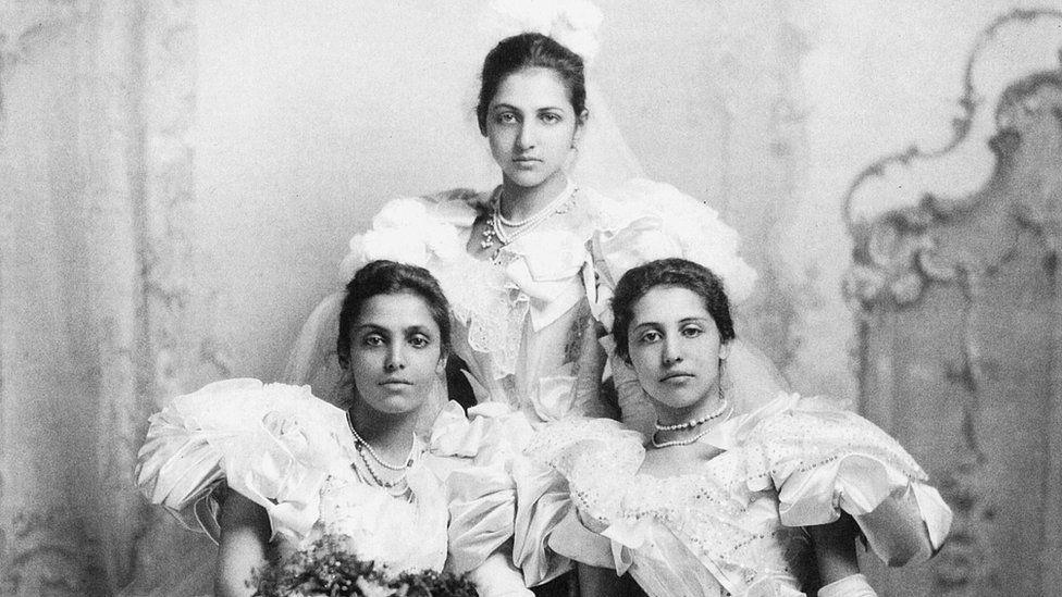 A black and white portrait of the three sisters. Two are sat side by side and one is sitting slightly elevated. They are wearing big dresses with a lot of frills and jewels. None of them are smiling but they are all looking at the camera.