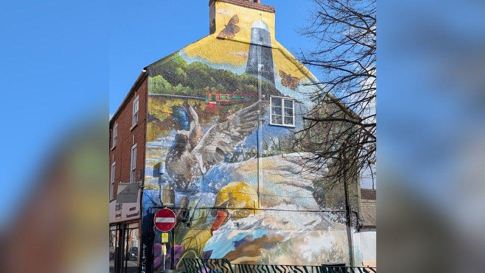 The colourful mural on the side of the building depicts the River Slea with a canal boat and mill. In the foreground is a duck and a swan splashing their wings in the water. There is a Treeline Dental sign to the side of the mural.