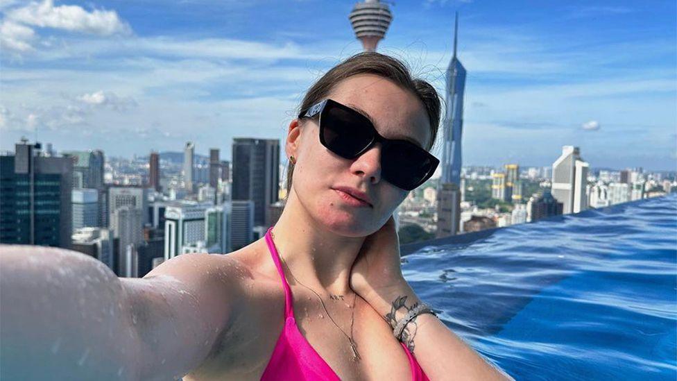 A selfie of Valeria Baigascina taken in a rooftop pool in Kuala Lumpur, with the striking skyline with tall towers behind her. Her long dark hair is tied back, she wears a pink bikini and dark sunglasses and a tattoo is visible on her wrist.