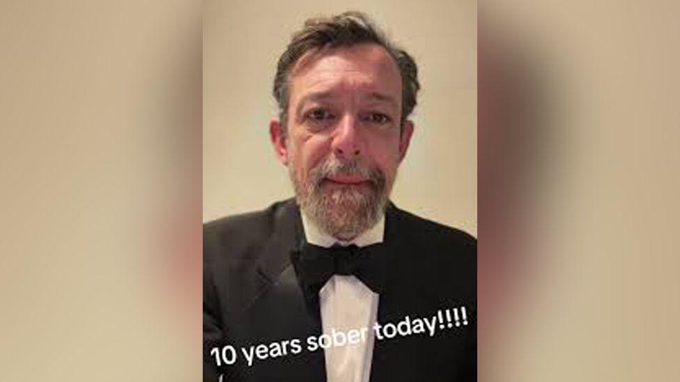 A still image from one of Justin Bone's TikTok videos. He is wearing a black tie suit and text reading "10 years sober today!!!!" is written as on-screen text in the lower quarter of the image.