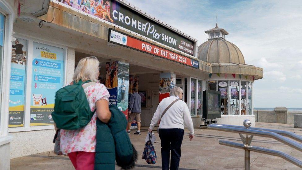Two women walking on Cromer Pier. They both have their backs to the camera and you cannot see their faces. On the pier is a sign which reads, CROMER Pier SHOW. PIER OF THE YEAR 2024.