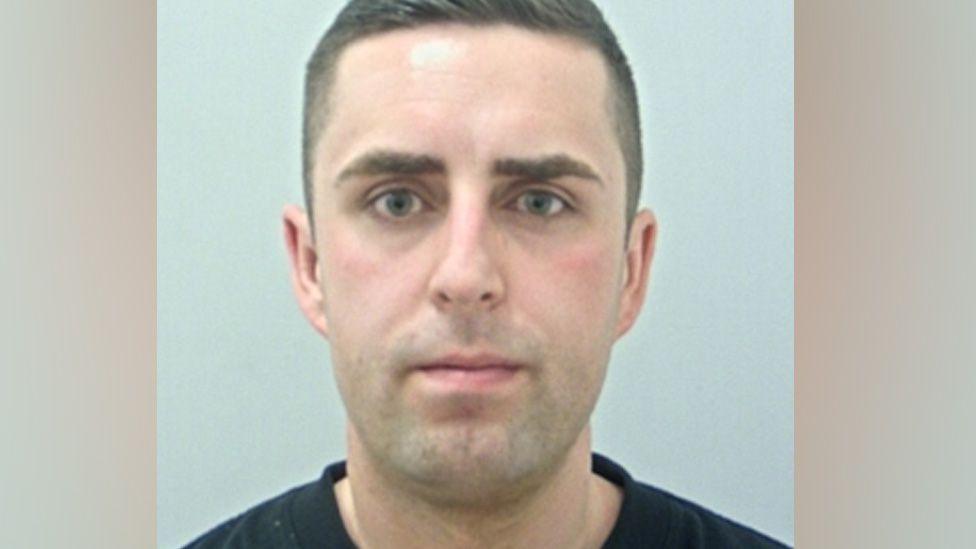 Joshua Adcroft's mugshot. He has short dark hair and is wearing a black t-shirt against a plain grey background