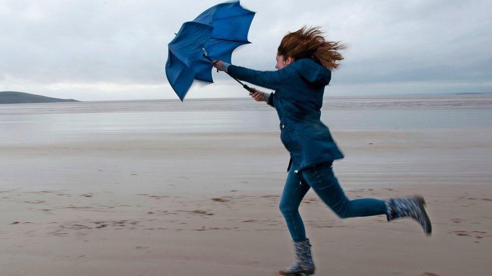 Woman with umbrella on a beach. She is wearing a blue jacket and jeans and the umbrella is blue too. She is being pulled along by heavy winds