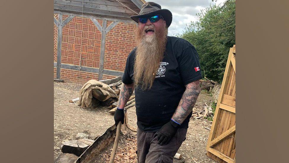 A man with a long beard, wearing a hat, holding a handmade chisel laughing