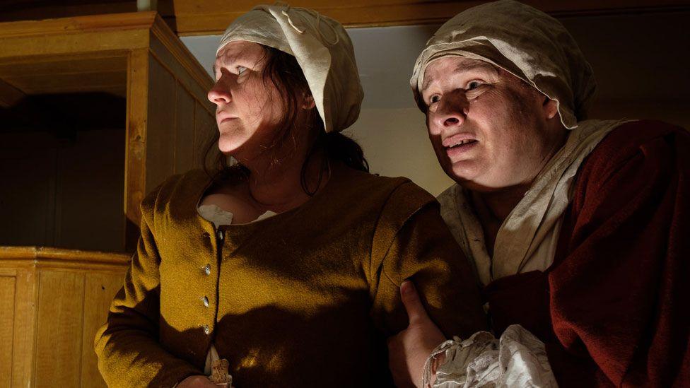 Two women in mid-17th Century dress during a re-enactment of the Huntingdon witch trial. They have white linen caps on their heads and are wearing russety-toned dresses of wool. They are looking frightened.