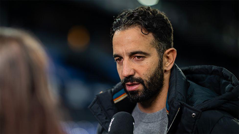 Ruben Amorim, speaking into a black microphone being held by a reporter. He is wearing a grey sweater and a large dark coloured rain coat with a hood.