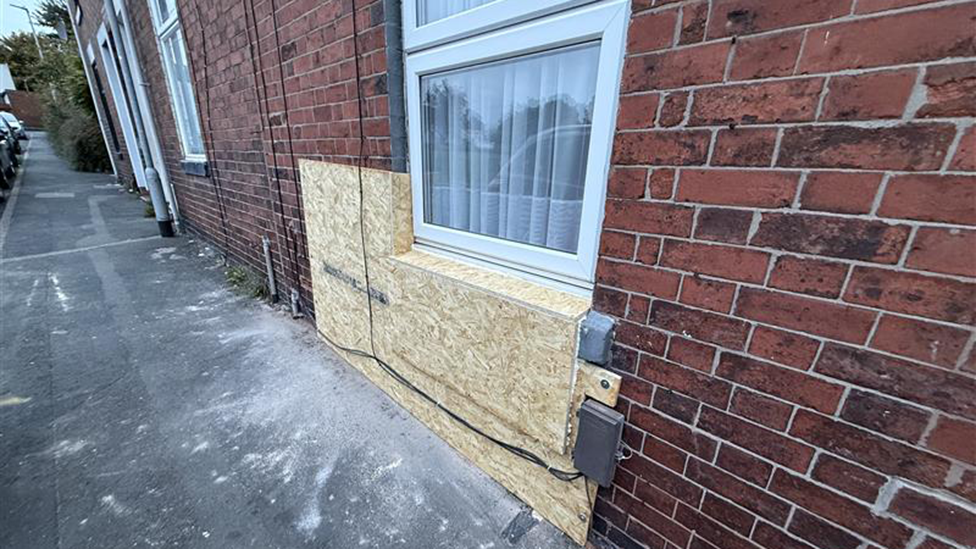 Part of a house, showing a section under a window boarded up, with the remains of debris underneath