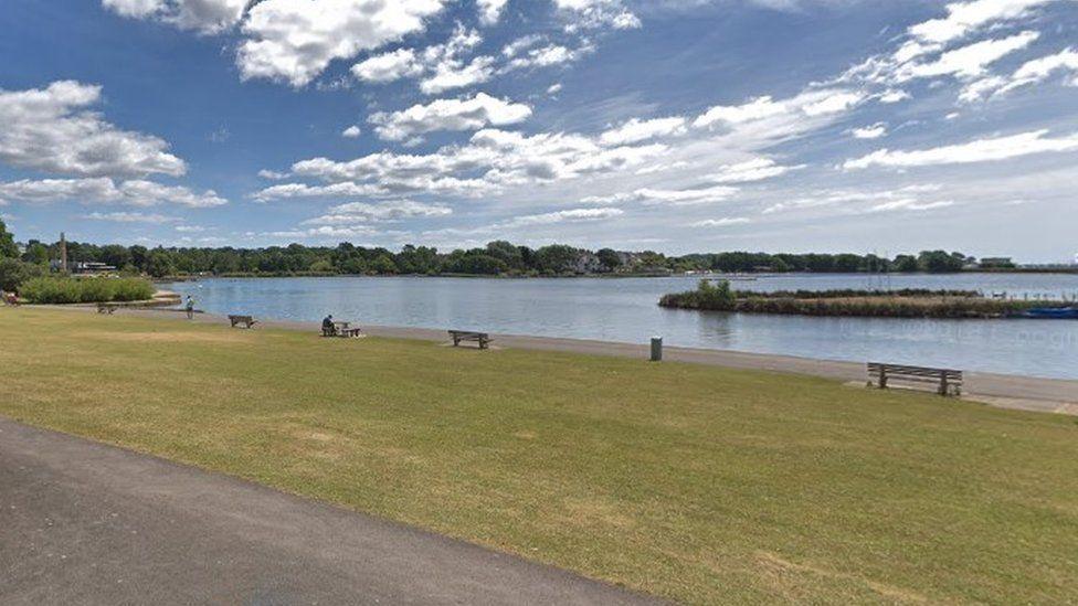 Grass area with path and benches along the edge of a large area of water with an island.