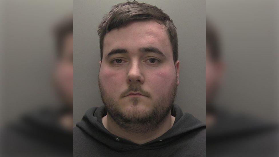 Custody photograph of Connor Laud he has short brown hair and beard and is wearing a dark hooded top