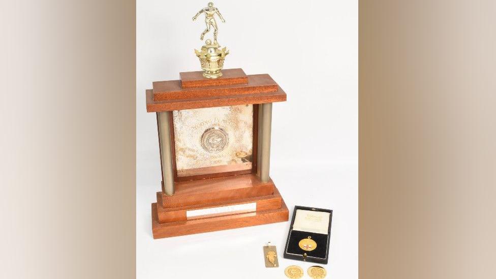 A large trophy made from light brown wood. It has a gold plaque in its middle and a gold statue of a footballer kicking a ball on top of it. Next to it, is a circular gold medal in a cushioned box, with two other gold circular medals next to it, and a silver rectangular medal.