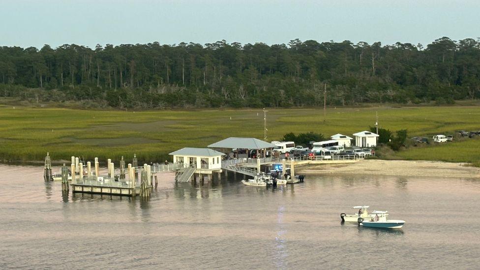 An aerial photo shows the Sapelo Island dock, which is formed of two small buildings and a jetty extending from the mainland into the sea, with a forest of trees in the background. The jetty is partially collapsed, and emergency service boats surround the dock with a crowd of people and traffic on the land side of the dock.