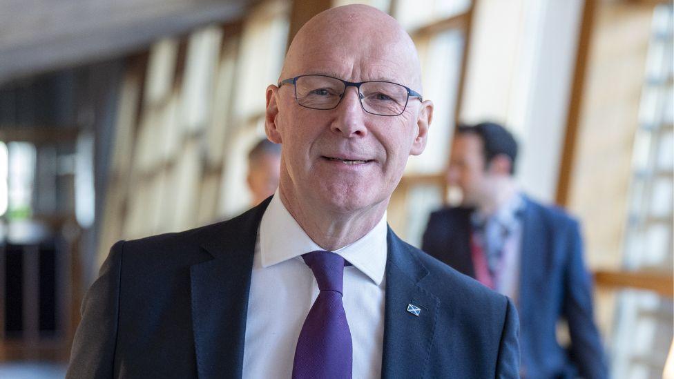 John Swinney, a bald man with glasses, smiles at the camera as he walks in the Scottish Parliament. He is visible from the chest up. He is wearing a dark suit, white shirt and purple tie, with a small saltire badge on his jacket. 