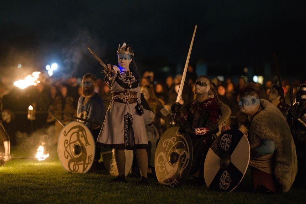 A woman dressed as a Celtic queen holding a sword out. Men dressed as Celtic warriors with blue and white face paint, shields and swords are crouched behind her. 