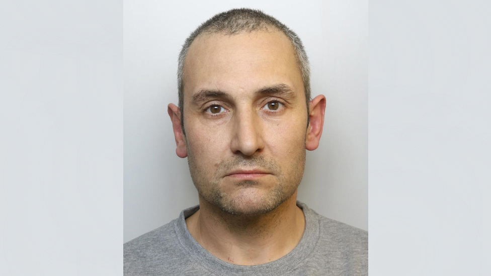 Mugshot of Antony Snook. He is wearing a grey T-shirt and standing against a grey background. He has short black and grey hair, brown eyes and thin lips, with a stern expression on his face.