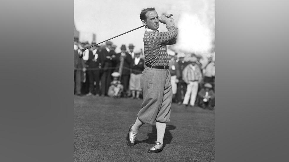 Old black and white image of golfer, Bobby Cruickshank, swinging golf club, with trousers tucked into socks, with crowd of smartly-dressed spectators in the background.