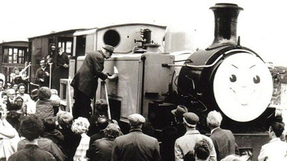 A black and white image showing the Reverend W Awdry standing on a step ladder and leaning towards the body of a steam engine.  The engine has a round face which has been painted with smiling features beneath a black steam chimney. Crowds of men and women are gathered around author. 