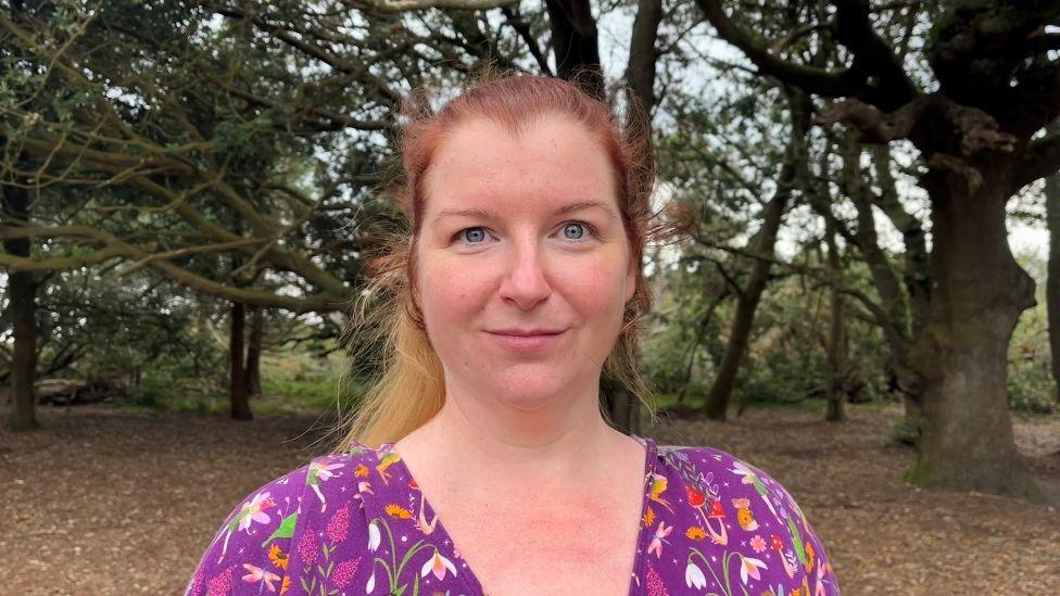 Charlotte Shenkin standing in a wooded area. She has long reddish-blonde hair pulled back, and she's wearing a purple shirt with colourful patterns of small plants and flowers, and mushrooms