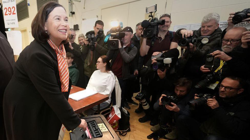 Mary Lou McDonald, wearing a dark suit jacket and orange and and white scarf places her ballot into a box while a crowd of photographers take pictures.