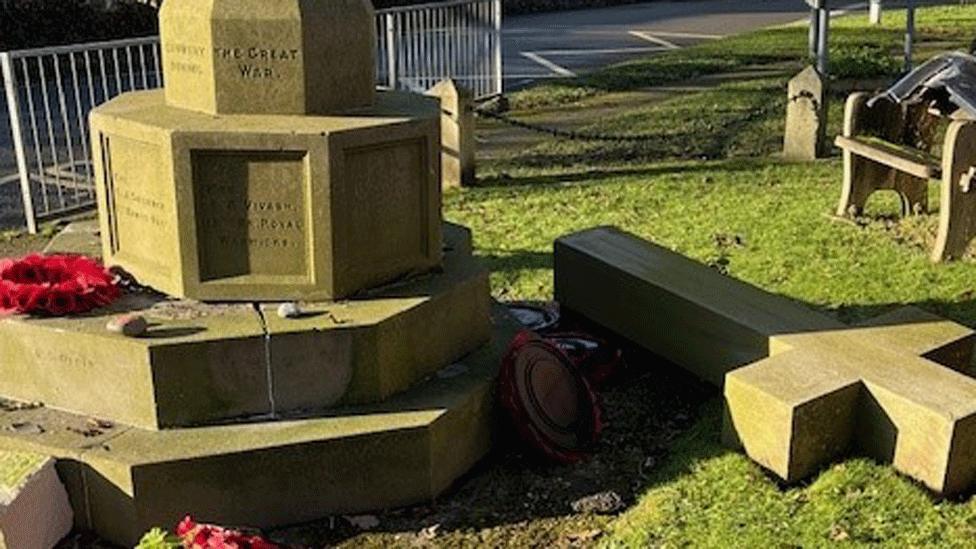 A war memorial is cracked and broken, its cross fallen onto the grass next to it. Wreaths and debris are scattered around.