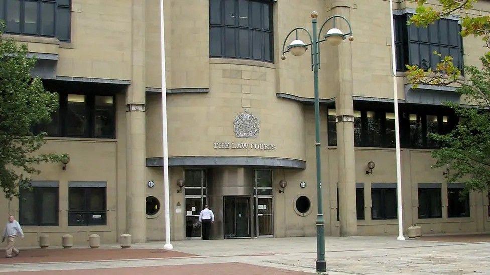 The front of Bradford Crown Court: a beige building with windows to the side and above the entrance. A lamp-post is in the foreground.