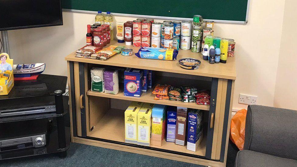 Packaged food lined up inside a cream wooden cupboard. There are tins, jars, cardboard packages and bags. There are also household goods, such as deodorant and body wash.