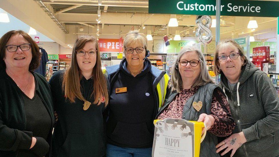 Five women stood in line under a customer service sign they all are wearing glasses and one holds a yellow fundraising bucket.
