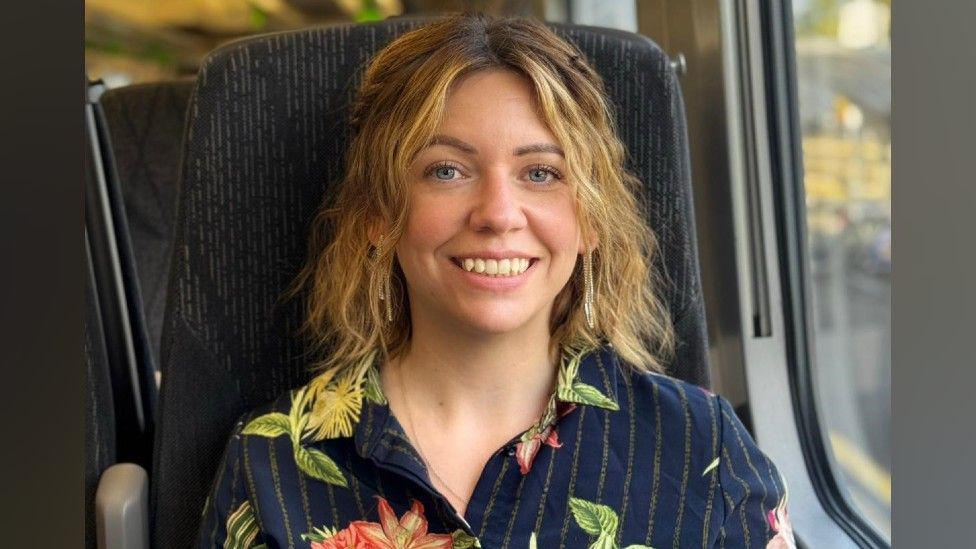 A woman with blonde wavy hair is sitting in a train seat, smiling at the camera. She is wearing a navy blouse with red and pink flowers and green leaves on it.