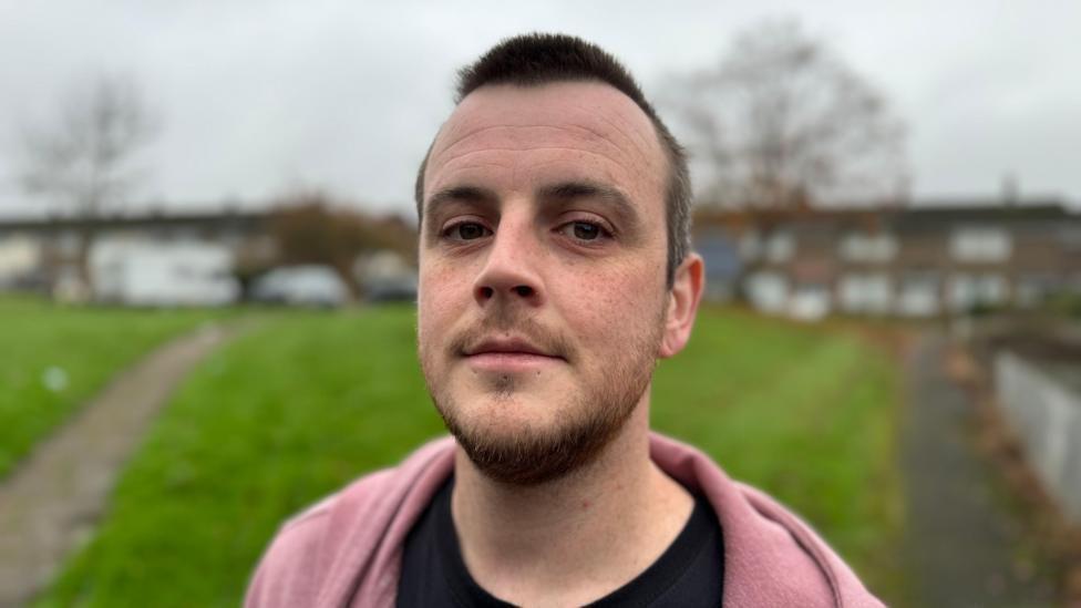 Marcus Johnson, 34, from Cwmbran, Torfaen, stands in front of some houses and a green.