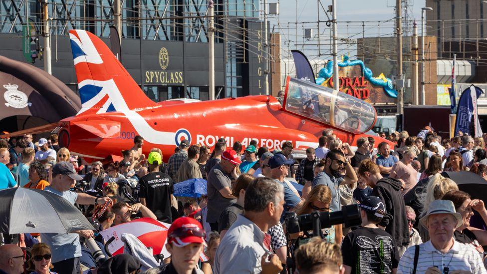 Dozens of people crowd around a red, white and blue Red Arrows plane, which has its cockpit open on Blackpool's Promenade