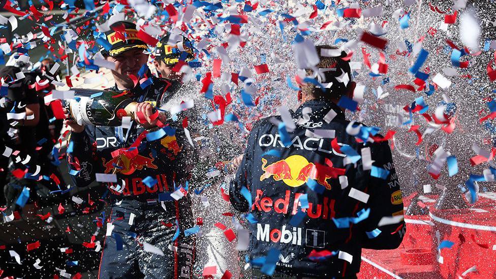 Red Bull Formula 1 drivers celebrate on the podium, spraying champagne as red, white, and blue confetti fills the air. Racing suits display Oracle and Mobil 1 logos.