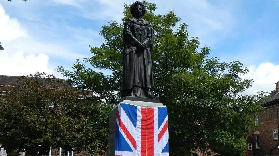 Union flag wrapped around the plinth of a bronze sculpture depicting Margaret Thatcher. The sculpture is surrounded by trees.