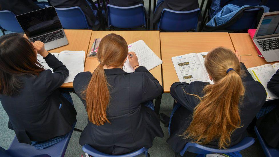 Three high school girls sit at a desk in a classroom. They have their backs to the camera. They are writing on tests papers.