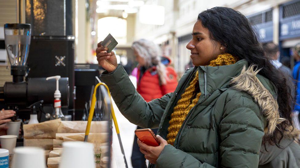 A woman wearing a green winter coat and yellow scarf pays for a coffee in a market