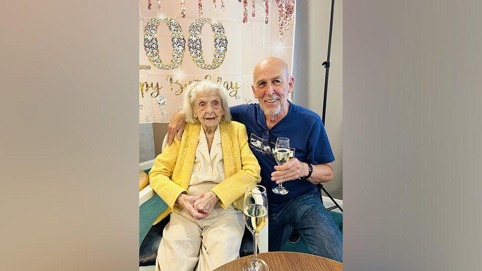 Elderly woman in a yellow blouse and jacket wears cream slacks is being given a hug by a man with greying hair and goatee beard. He is smiling and holds a glass of champagne. Another glass of champagne is on a table in front of them.