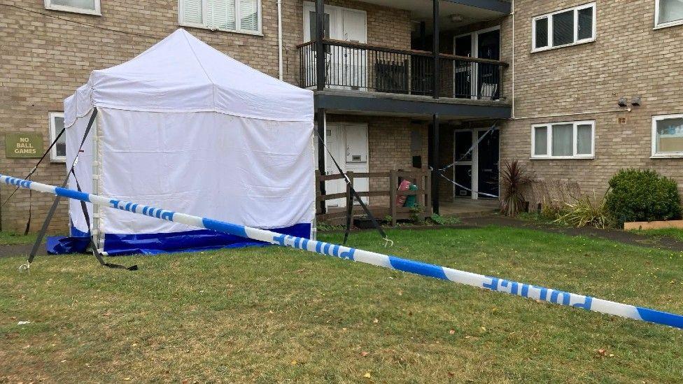 A block of flats with a white police tent outside and blue and white police cordon tape.