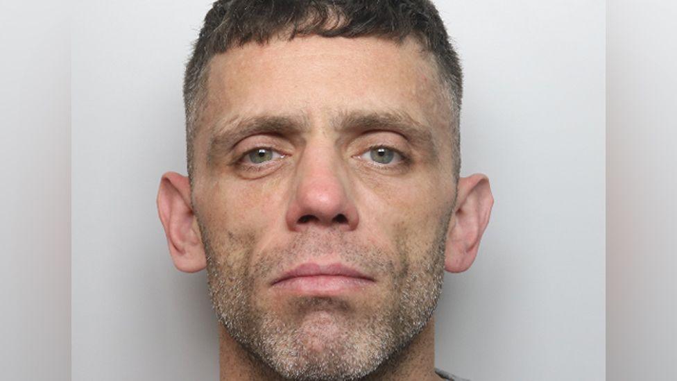 A police mug shot of Shaun Roadnight, he is looking straight to camera, has grey stubble and short dark hair.