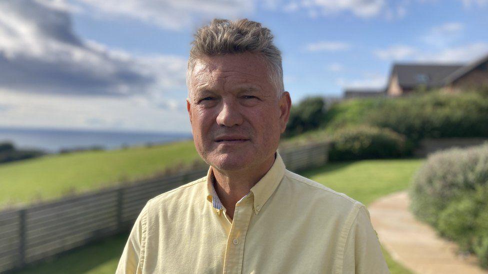 A photo of Mike Starkie who has grey short hair and is wearing a yellow shirt. 