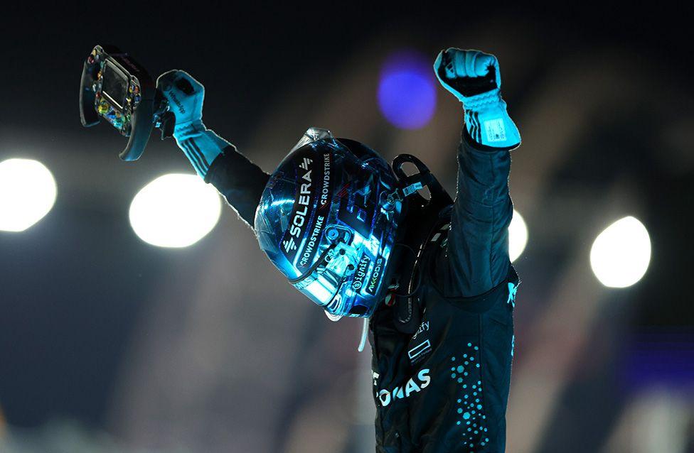 Race winner George Russell celebrates during the F1 Grand Prix of Singapore at the Marina Bay Street Circuit. The image shows Russell in a black racing suit and reflective helmet, raising both arms in victory while holding a steering wheel, with bright lights illuminating the night background.