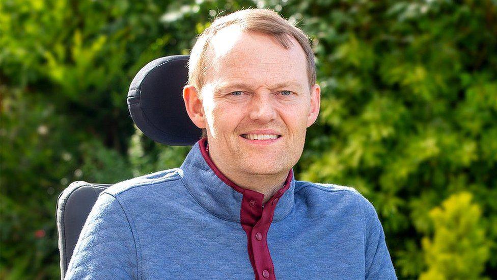 Scott Stewart smiles at the camera while sitting outside in his wheelchair in the sunlight with bushes in the background