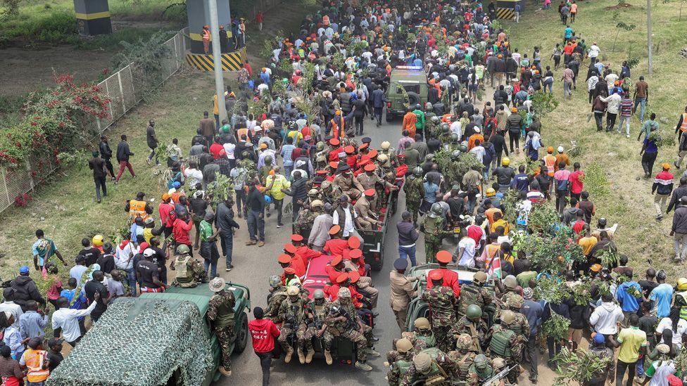 Birdseye view of the funeral cortege for Raila Odinga, showing the vehicle carrying the coffin surrounded by officers some dressed in red - and crowds thronging around it.