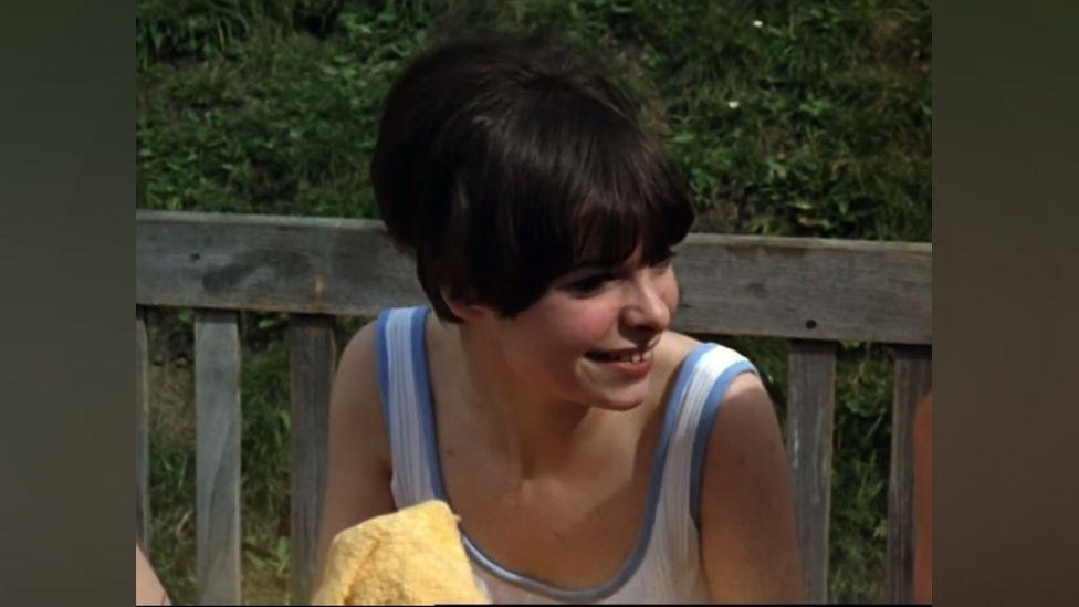 Seventeen-year-old Maggie Hackney in the film "The Givers". She sits on a wooden bench outdoors, with a hedge behind. She wears a casual vest top which is white with pale blue edging. Her hair is dark brown and short, in a typical 1960s bouffant