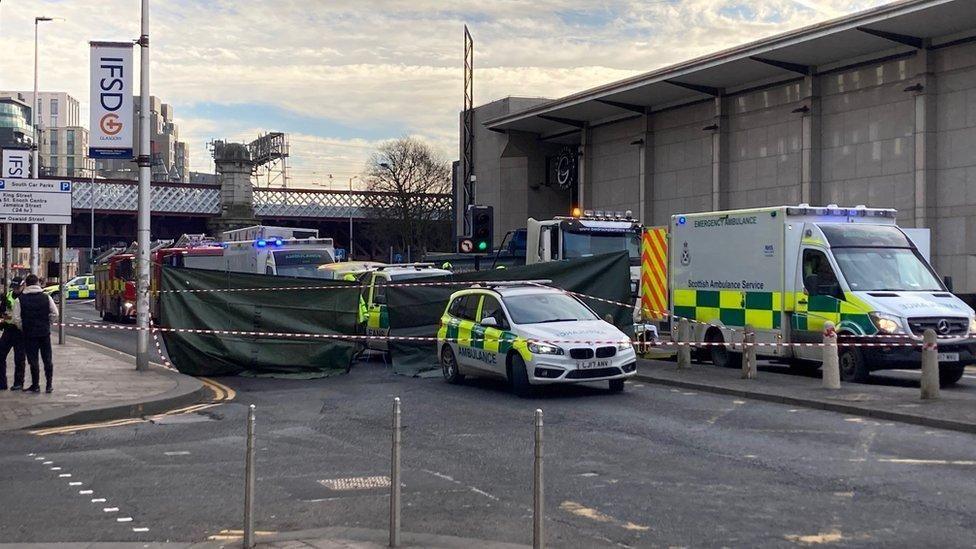 Emergency service vehicles at the scene of the accident, which is cordoned off with tape. Several emergency service vehicles are on the scene, which an ambulance and a marked car in the foreground. Other vehicles are partially visible behind two large dark green sheets, which are acting as a cordon at the scene. Off to the left a police officer appears to be interviewing a witness on the pavement.