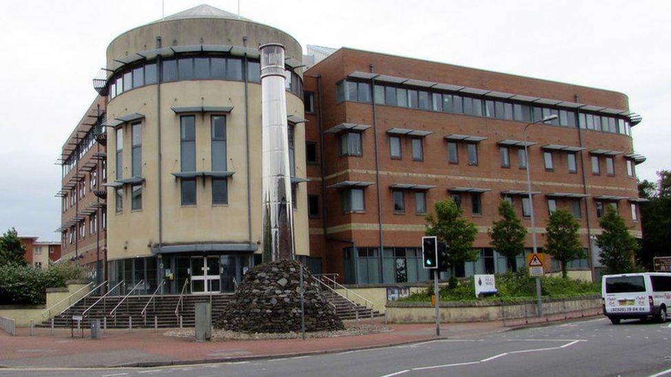 The outside of a police station building, which is brown and white with lots of windows and stairs at the front. In front of the building is a main road, with traffic lights.